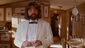 Movie still from “Masterminds” (2015), directed by Jared Hess – A man in a white tuxedo holding a piece of food; Medium shot, Over the shoulder angle