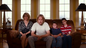 Movie still from “Masterminds” (2015), directed by Jared Hess – A man sitting on top of a brown couch next to two women and a boy; Medium shot, High angle