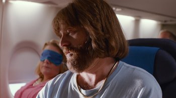 Movie still from “Masterminds” (2015), directed by Jared Hess – A man with long hair and a beard wearing a necklace; Medium shot, Over the shoulder angle