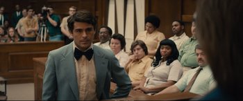 Movie still from “Extremely Wicked, Shockingly Evil and Vile” (2019), directed by Joe Berlinger – A man in a suit standing in front of a group of people in a court room; Medium shot, Over the shoulder angle