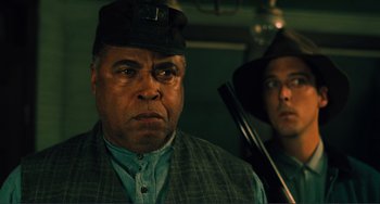 Movie still from “Matewan” (1987), directed by John Sayles – A man in a hat is looking at the camera while another man is holding a rifle; Close Up shot, Low angle