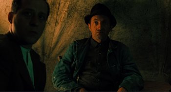 Movie still from “Matewan” (1987), directed by John Sayles – A man wearing a hat sitting in front of a wall; Medium shot, Low angle