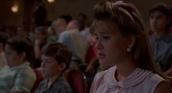 Movie still from “Matinee” (1993), directed by Joe Dante – A girl with a bow in her hair sitting in front of a crowd; Close Up shot, Over the shoulder angle