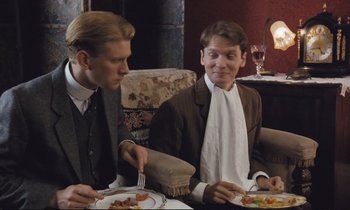 Movie still from “Maurice” (1987), directed by James Ivory – Two men sitting at a dinner table with plates of food in front of them; Medium shot, Over the shoulder angle
