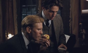 Movie still from “Maurice” (1987), directed by James Ivory – Two men in suits eating a donut in front of a window; Medium shot, Over the shoulder angle