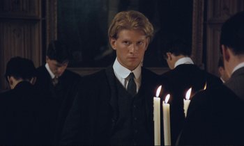 Movie still from “Maurice” (1987), directed by James Ivory – A man in a suit standing in front of a group of candles; Medium shot, Low angle