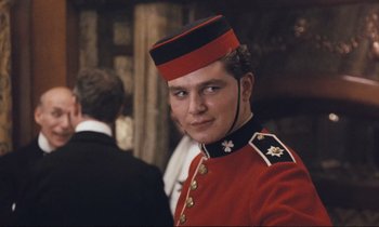 Movie still from “Maurice” (1987), directed by James Ivory – A man in a red and black uniform is posing for a picture; Close Up shot, Over the shoulder angle
