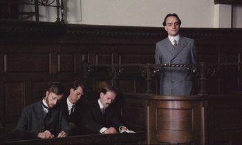 Movie still from “Maurice” (1987), directed by James Ivory – A man in a gray suit stands at a podium while four other men look on; Medium shot, Low angle