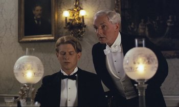 Movie still from “Maurice” (1987), directed by James Ivory – Two men in tuxedos stand next to each other in front of lamps; Medium shot, Low angle