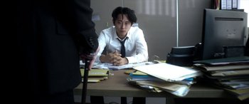 Movie still from “Mayhem” (2017), directed by Joe Lynch – A man sitting at a desk in front of papers; Medium shot, Over the shoulder angle