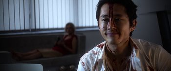 Movie still from “Mayhem” (2017), directed by Joe Lynch – A young man with blood all over his face and a woman sitting in a chair in the background; Close Up shot, Over the shoulder angle
