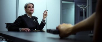 Movie still from “Mayhem” (2017), directed by Joe Lynch – A woman sitting at a table holding a cigarette in her hand; Close Up shot, Over the shoulder angle