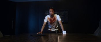 Movie still from “Mayhem” (2017), directed by Joe Lynch – A man with blood all over his face is sitting at a table; Medium shot, Low angle