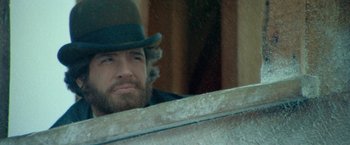 Movie still from “McCabe & Mrs. Miller” (1971), directed by Robert Altman – A man with a beard and a green hat is leaning on a wall; Close Up shot, Over the shoulder angle