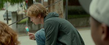 Movie still from “Me and Earl and the Dying Girl” (2015), directed by Alfonso Gomez-Rejon – A man sitting on the ground looking at his cell phone; Medium shot, Over the shoulder angle