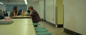 Movie still from “Me and Earl and the Dying Girl” (2015), directed by Alfonso Gomez-Rejon – A man sitting at a table in front of a row of blue stools; Wide shot, Over the shoulder angle
