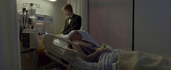 Movie still from “Me and Earl and the Dying Girl” (2015), directed by Alfonso Gomez-Rejon – A bald man in a suit is laying in a hospital bed; Medium shot, Over the shoulder angle