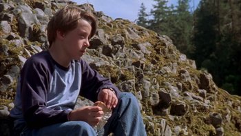 Movie still from “Mean Creek” (2004), directed by Jacob Estes – A boy sitting on the ground in front of a rock wall; Medium shot, Over the shoulder angle