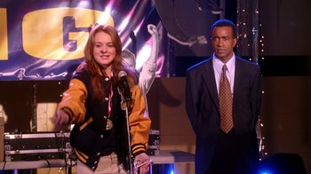 Movie still from “Mean Girls” (2004), directed by Mark Waters – A woman standing in front of a microphone next to a man in a suit; Medium shot, Over the shoulder angle