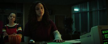 Movie still from “Fear Street: Part One - 1994” (2021), directed by Leigh Janiak – A woman sitting at a table in a dark room; Medium shot, Low angle