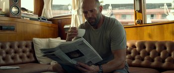 Movie still from “Mechanic: Resurrection” (2016), directed by Dennis Gansel – A man sitting on a bed holding a coffee cup and reading a newspaper; Close Up shot, Over the shoulder angle