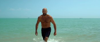 Movie still from “Mechanic: Resurrection” (2016), directed by Dennis Gansel – A man walking through the water on the beach; Medium shot, Low angle