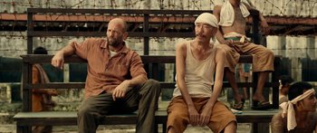 Movie still from “Mechanic: Resurrection” (2016), directed by Dennis Gansel – Two men sitting on a wooden bench in front of a fence; Medium shot, Over the shoulder angle