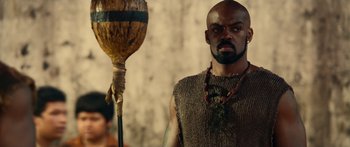 Movie still from “Mechanic: Resurrection” (2016), directed by Dennis Gansel – A man with a beard is standing next to an object; Medium shot, Low angle