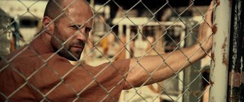 Movie still from “Mechanic: Resurrection” (2016), directed by Dennis Gansel – A bald man is behind a chain link fence; Close Up shot, Over the shoulder angle