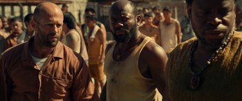 Movie still from “Mechanic: Resurrection” (2016), directed by Dennis Gansel – A man with a beard is standing in front of a group of people; Medium shot, Over the shoulder angle