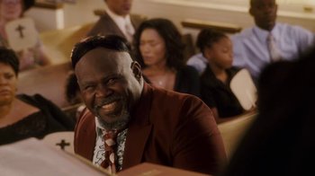 Movie still from “Meet the Browns” (2008), directed by Tyler Perry – A man in a red jacket smiles at the camera while people in the background look on; Close Up shot, Over the shoulder angle