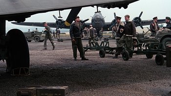 Movie still from “Memphis Belle” (1990), directed by Michael Caton-Jones – A group of men standing next to an airplane on the ground; Wide shot, Low angle
