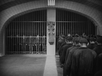 Movie still from “Metropolis” (1927), directed by Fritz Lang – Black and white photograph of men standing in front of bars; Wide shot, High angle