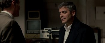 Movie still from “Michael Clayton” (2007), directed by Tony Gilroy – A man standing in a room next to a microphone; Close Up shot, Over the shoulder angle