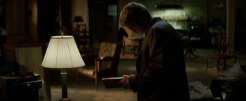 Movie still from “Michael Clayton” (2007), directed by Tony Gilroy – A man standing in a living room holding a book; Medium shot, Over the shoulder angle