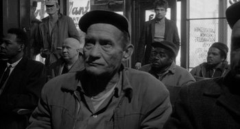 Movie still from “Mickey One” (1965), directed by Arthur Penn – An old man with a hat on and several other men in the background; Medium shot, Over the shoulder angle