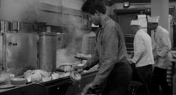 Movie still from “Mickey One” (1965), directed by Arthur Penn – A black and white photo of a man cooking in a kitchen; Medium shot, High angle
