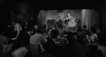Movie still from “Mickey One” (1965), directed by Arthur Penn – A group of people sitting at a table in front of a screen; Wide shot, Over the shoulder angle