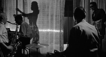 Movie still from “Mickey One” (1965), directed by Arthur Penn – A man and a woman playing drums in front of a glass wall; Medium shot, Over the shoulder angle