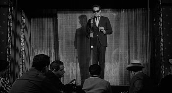 Movie still from “Mickey One” (1965), directed by Arthur Penn – A black and white photo of a man on a stage; Wide shot, Over the shoulder angle