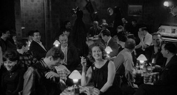 Movie still from “Mickey One” (1965), directed by Arthur Penn – A woman sitting at a table with a cigarette in her mouth; Medium shot, Over the shoulder angle