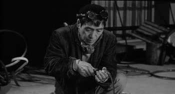 Movie still from “Mickey One” (1965), directed by Arthur Penn – An old man wearing goggles and a hat is working on something; Medium shot, High angle