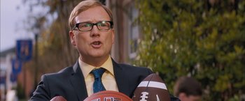 Movie still from “Middle School: The Worst Years of My Life” (2016), directed by Steve Carr – A man in a suit and tie holding a football and a basketball; Close Up shot, Over the shoulder angle