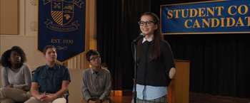 Movie still from “Middle School: The Worst Years of My Life” (2016), directed by Steve Carr – A woman standing in front of a microphone while a boy looks on; Medium shot, Over the shoulder angle