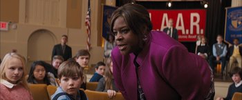 Movie still from “Middle School: The Worst Years of My Life” (2016), directed by Steve Carr – A woman in a purple jacket sitting in front of a group of people; Medium shot, Over the shoulder angle