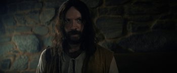 Movie still from “Fear Street: Part Three - 1666” (2021), directed by Leigh Janiak – A man with long hair and beard standing in front of a stone wall; Close Up shot, Over the shoulder angle