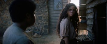 Movie still from “Fear Street: Part Three - 1666” (2021), directed by Leigh Janiak – A woman with long curly hair standing in front of a stone building; Medium shot, Over the shoulder angle