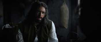 Movie still from “Fear Street: Part Three - 1666” (2021), directed by Leigh Janiak – A man with long hair and a beard is sitting; Medium shot, Over the shoulder angle