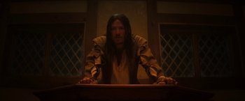 Movie still from “Fear Street: Part Three - 1666” (2021), directed by Leigh Janiak – A man with long brown hair standing at a table; Medium shot, Low angle