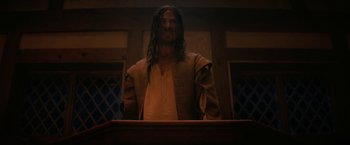 Movie still from “Fear Street: Part Three - 1666” (2021), directed by Leigh Janiak – A man with long dreadlocks standing in front of a podium; Medium shot, Low angle